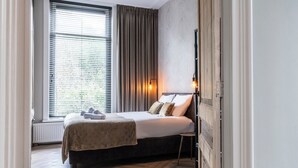 Un dormitorio con una cama, una mesita de noche, una ventana con persianas y una lámpara fijada a la pared.