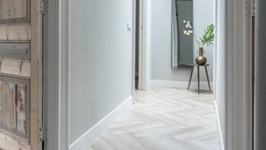 Un pasillo con una puerta de madera, una pared blanca, un espejo y una planta sobre un pequeño pedestal.