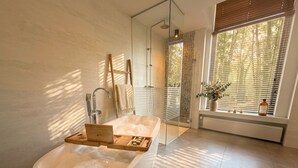 A modern bathroom with a freestanding bathtub, a glass shower enclosure, and a window with blinds. - First-floor suite, 35m2, ensuite bathroom with bath and rain shower. (Tilburg)