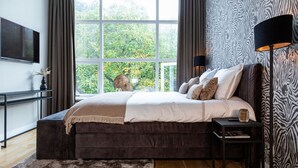 A bedroom with a large window, a bed, a bedside table, a lamp, and a TV. - First-floor suite, 35m2, ensuite bathroom with bath and rain shower. (Tilburg)