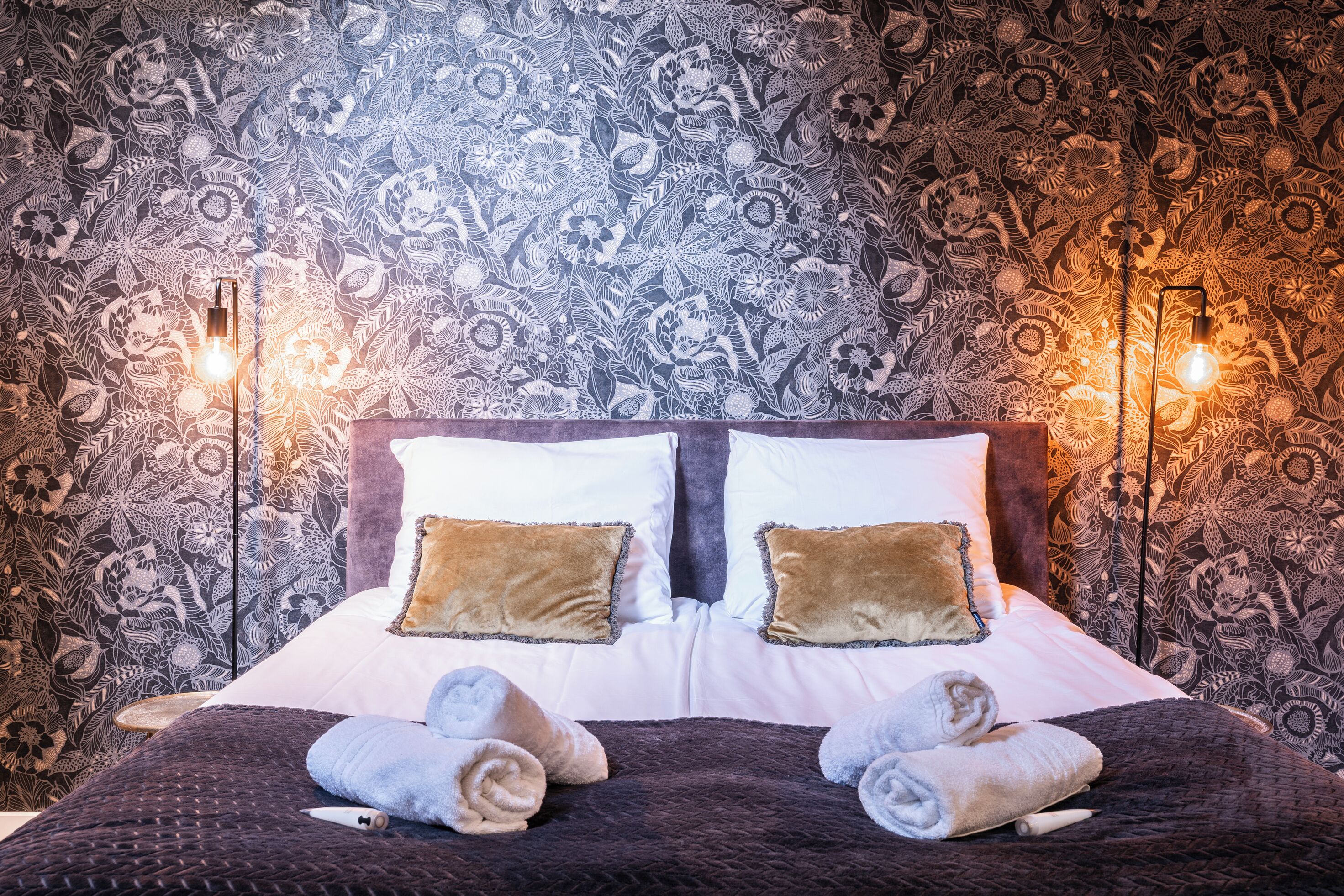 A bed with a patterned wallpaper, two bedside lamps, and two pillows.