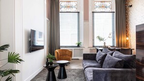 Una sala de estar moderna con un sofá gris, una mesita redonda, un televisor montado en la pared y dos ventanas con cortinas.