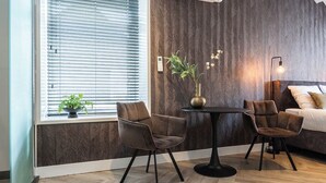 Una habitación moderna con una pared de madera en patrón espiga, una mesa redonda negra con dos sillas, una cama con ropa blanca y una ventana con persianas.