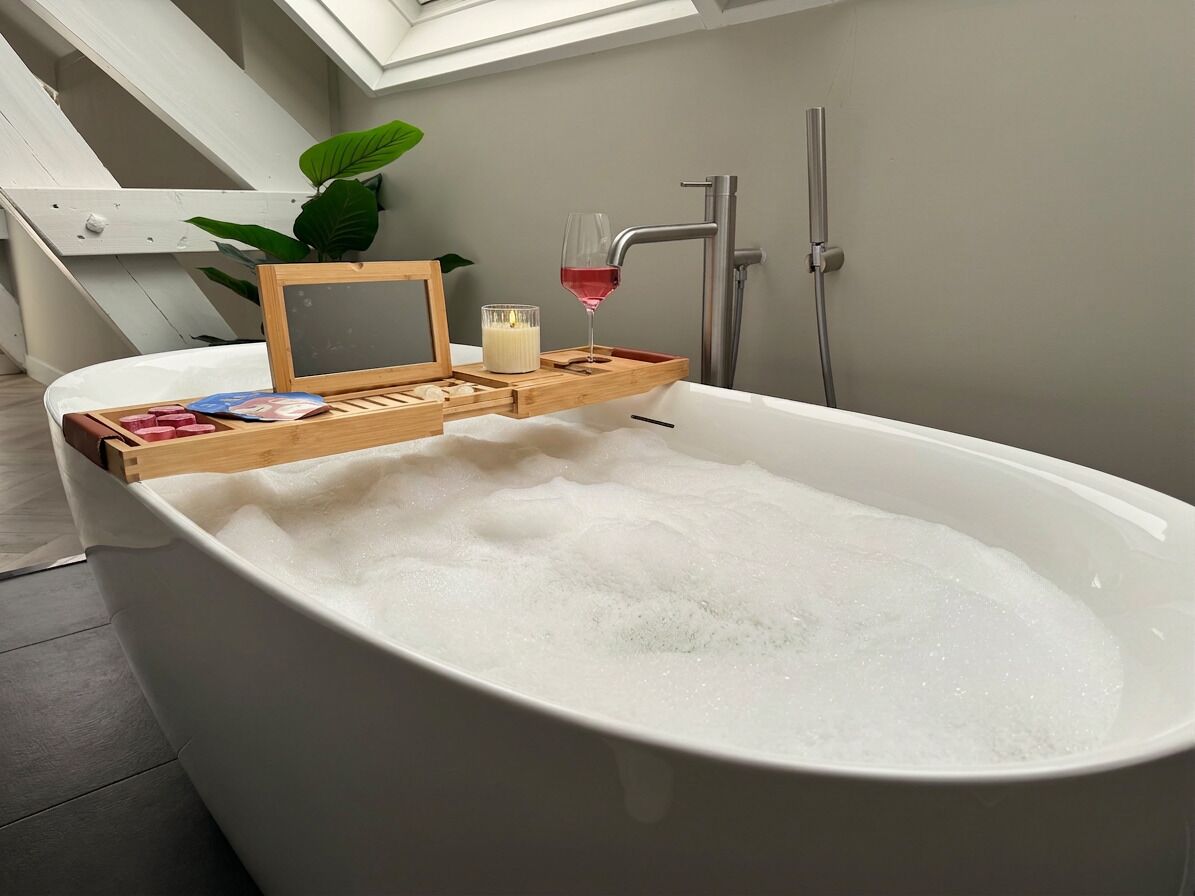 A bathtub with a wooden tray holding a glass of red wine and a candle, a plant, and a framed picture.