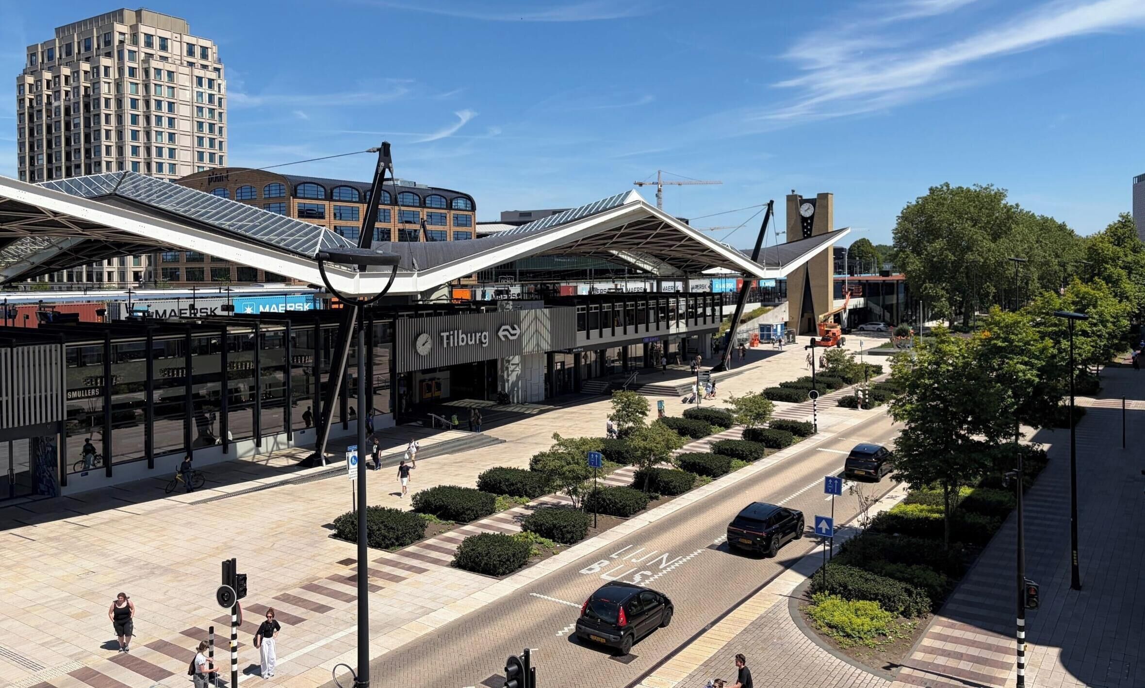 A modern train station with a large glass canopy, a clock tower, and a pedestrian crossing.