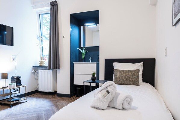 A hotel room with a bed, a desk, a chair, a small table, a radiator, a window with curtains, a mirror, and a TV mounted on the wall.