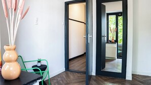 Un pasillo con piso de madera, una silla verde, un jarrón beige con cañas, y una puerta que da a una habitación con ventana.