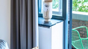 Una habitación con vistas al exterior, una ventana con cortina, un florero blanco con planta y una silla verde.