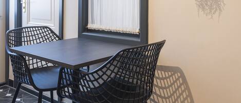 A room with a dark wooden table, two black chairs, a potted plant on the wall, and a window with white curtains.