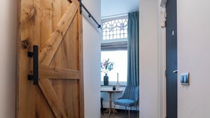 Un pasillo con una puerta de madera estilo granero, una ventana con cortinas, una silla y una pequeña mesa con una planta.