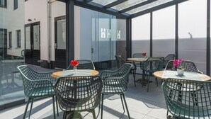 Un área de estar moderna al aire libre con paredes de vidrio, sillas y mesas de metal, con vistas a un paisaje urbano.