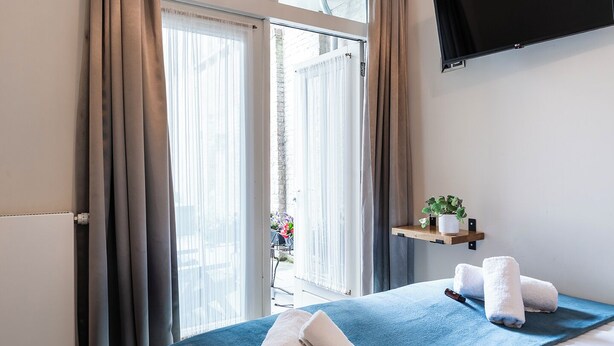Habitación de hotel con una cama, un televisor montado en la pared, una ventana con cortinas y un estante pequeño con una planta.