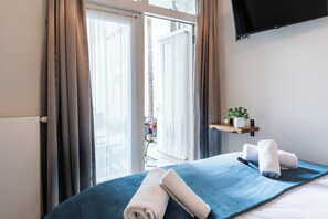A hotel room with a bed, a TV mounted on the wall, a window with curtains, and a small shelf with a plant. - Stay in the center of Tilburg with fast Wi-Fi and an easy check-in (Tilburg)