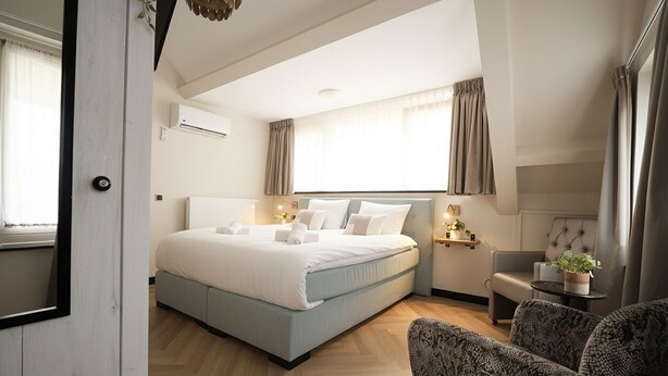 A modern hotel room with a large bed, a small seating area, and a window with curtains.