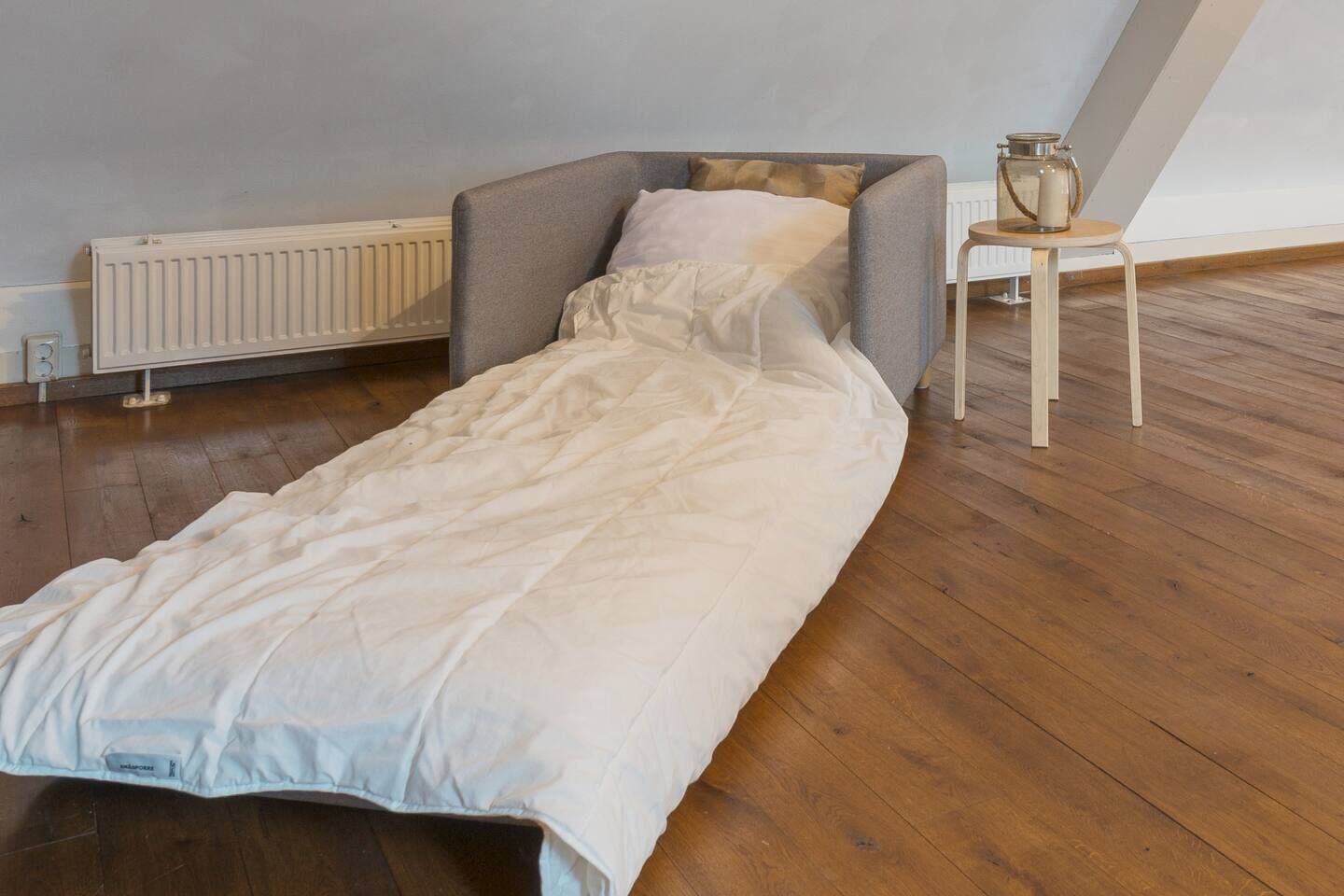 A single bed in a room with wooden flooring, a radiator, and a small table with a container.