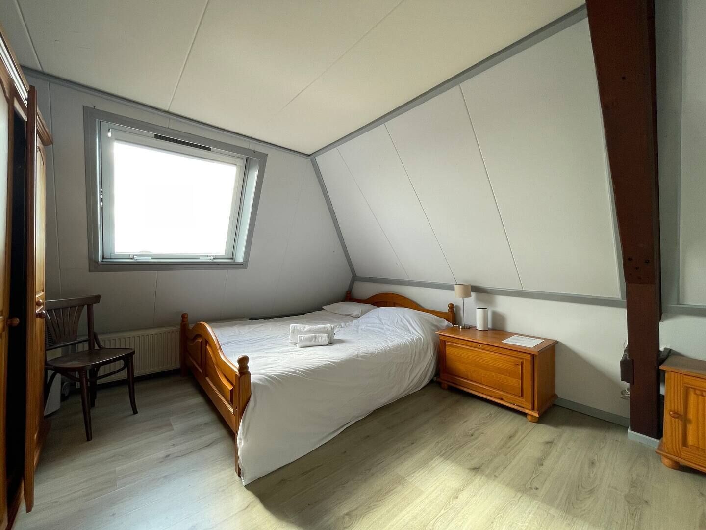 Une petite chambre bien éclairée, avec un lit simple, une table de chevet en bois, une chaise et une fenêtre laissant entrer la lumière naturelle.