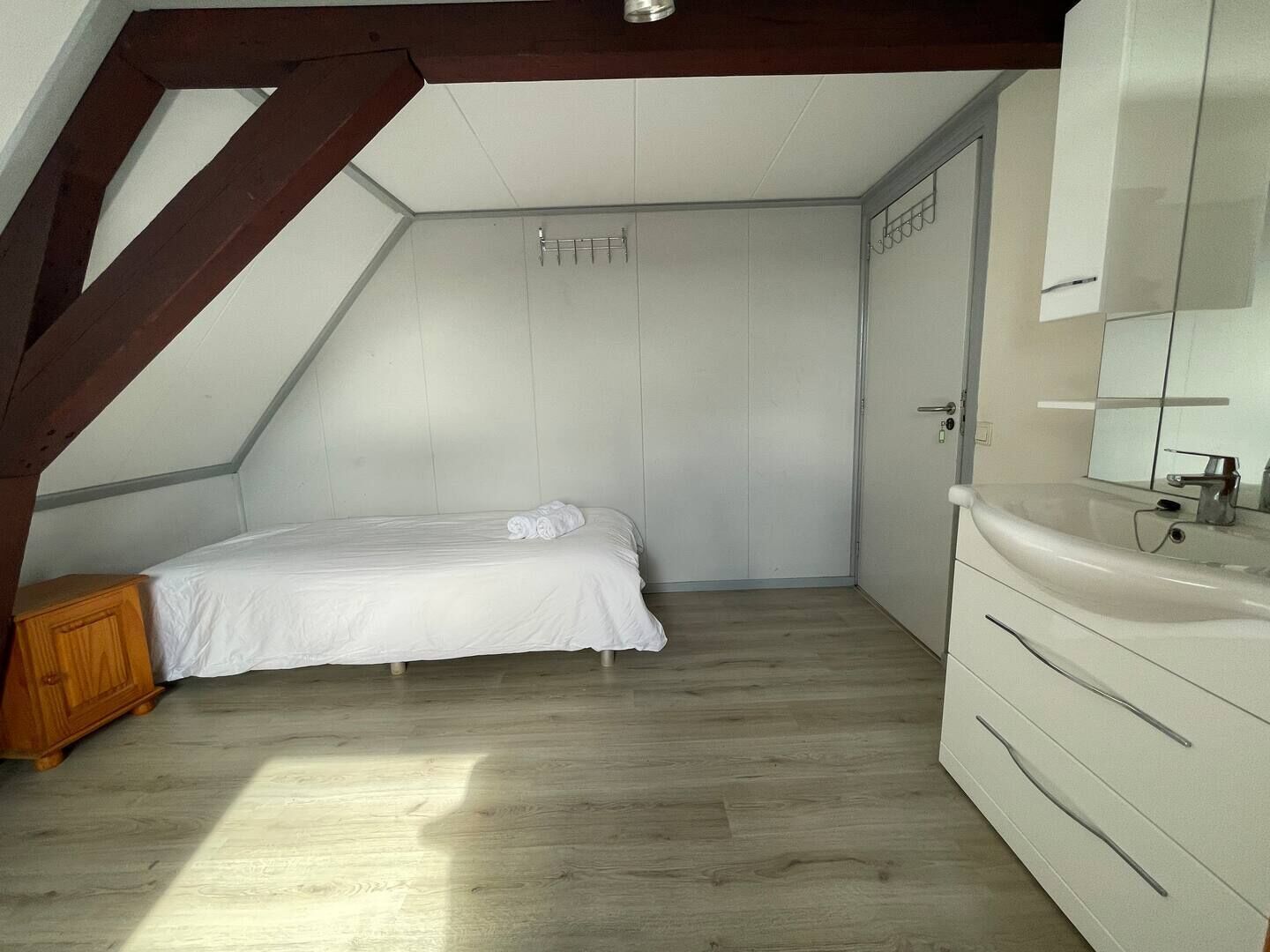 Un dormitorio con techo inclinado, una cama, un armario de madera, un tocador blanco y un baño con lavamanos y espejo.