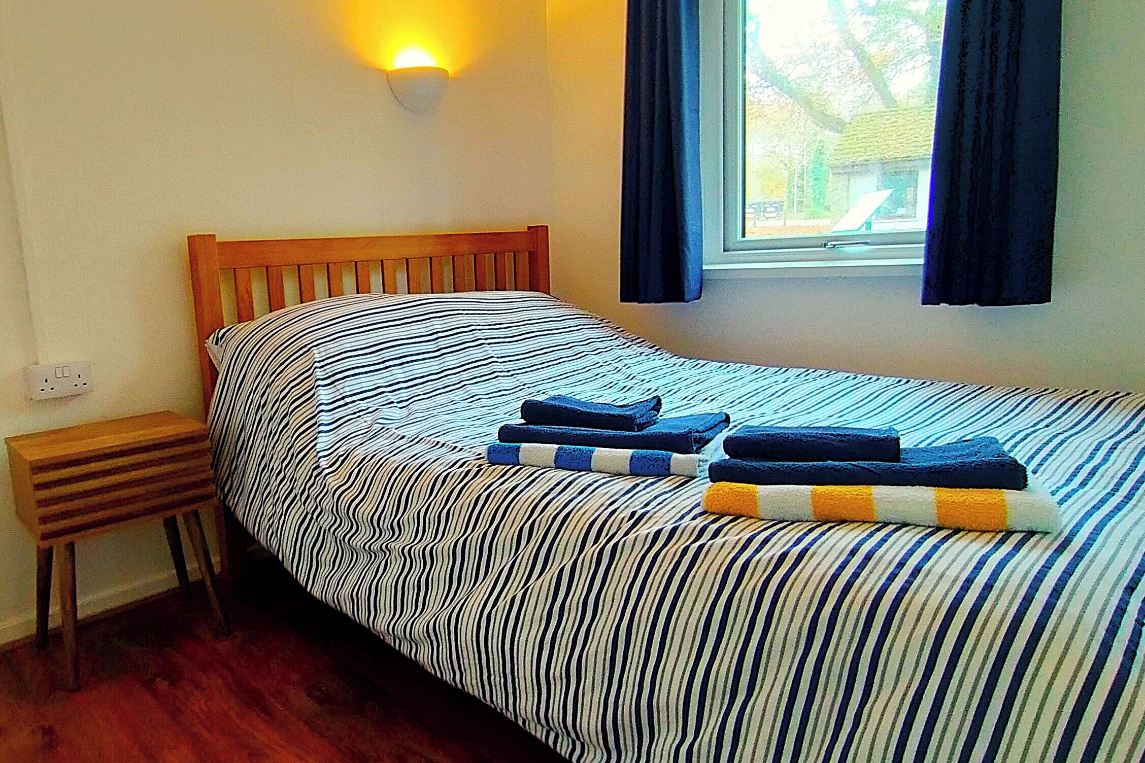 Una cama individual con ropa de cama a rayas, un cabecero de madera, una mesita de noche y una ventana con cortinas.