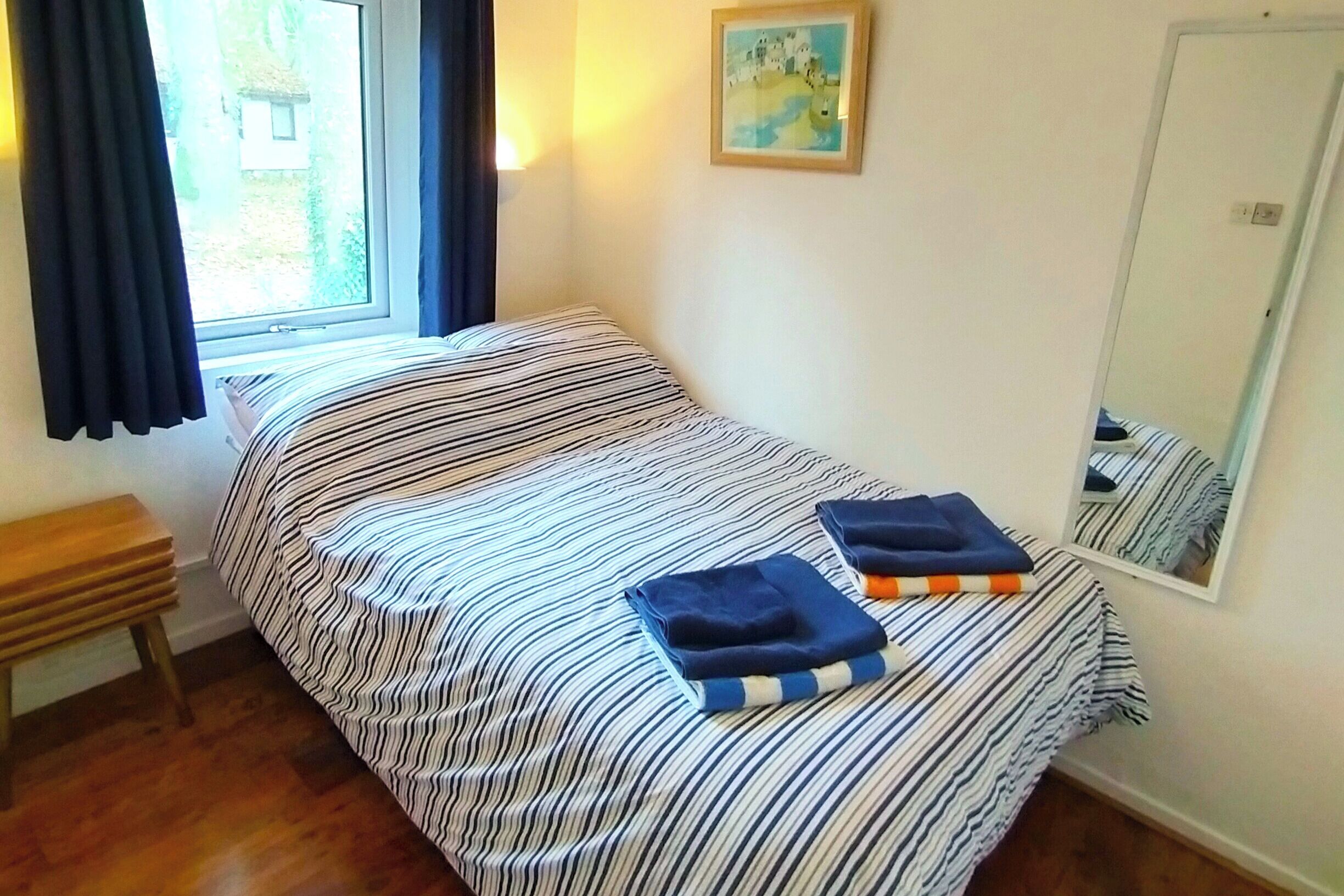 A bedroom with a bed, a bedside table, a window with curtains, a painting on the wall, and a mirror.