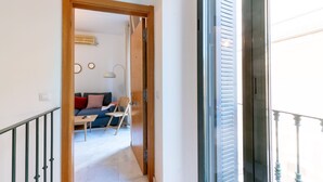 A hallway leading to a living room with a sofa, a table, and a chair. There is a door with a number plate, and a window with blinds. - Dos de mayo - One Bedroom Apartment, Sleeps 3 (Sevilla)