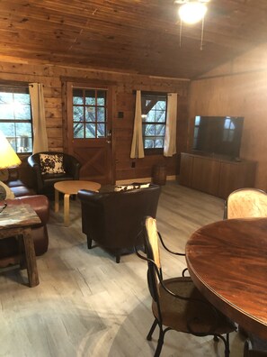 A cozy cabin interior with a wooden floor, leather furniture, a television, and large windows with curtains. - Cedar Ridge Log Cabin on 14 acres - Pet Friendly! (Granbury)