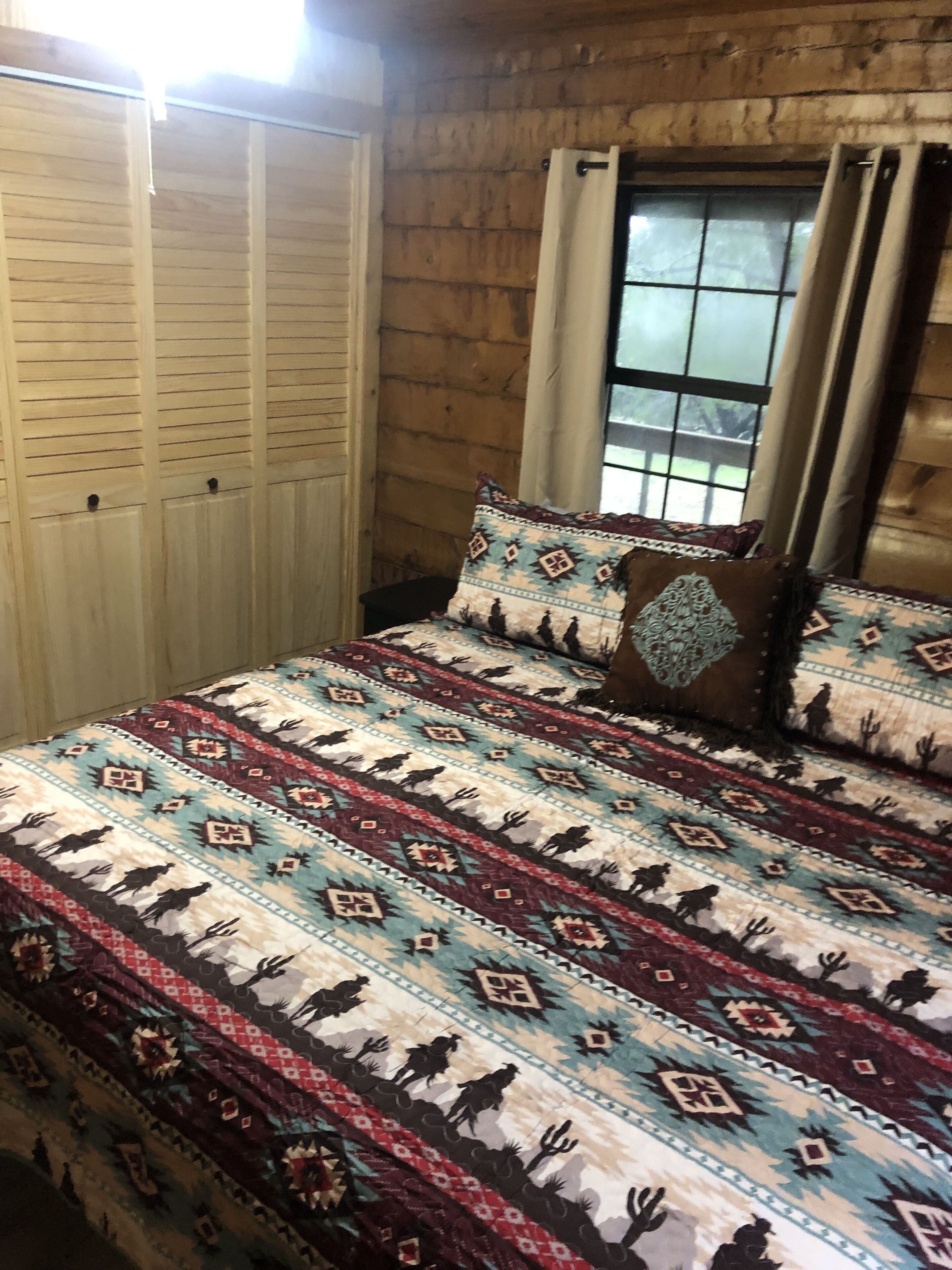 A room with a bed featuring a patterned bedspread, wooden walls, a window with curtains, and a door with a handle.