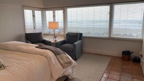 A bedroom with a bed, two armchairs, a small table with a lamp, and large windows with blinds. - Stunning Oceanfront Home with Private Beach Access - 468 Tarpon (Fripp Island)