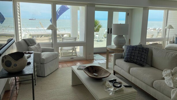 A spacious living room with a large window overlooking a beach and ocean. There is a white sofa, a coffee table, and a dining area with chairs. - Stunning Oceanfront Home with Private Beach Access - 468 Tarpon (Fripp Island)
