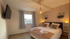Un dormitorio con cama, televisor montado en la pared, ventana con cortinas y lámpara en el techo.