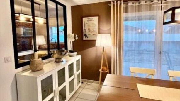 Una cocina moderna con zona de comedor, que presenta un amplio ventanal con cortinas, un armario blanco con puertas de vidrio y una mesa de comedor de madera.