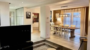 Una sala moderna con televisor de pantalla plana, un comedor con mesa y sillas, y una cocina al fondo.