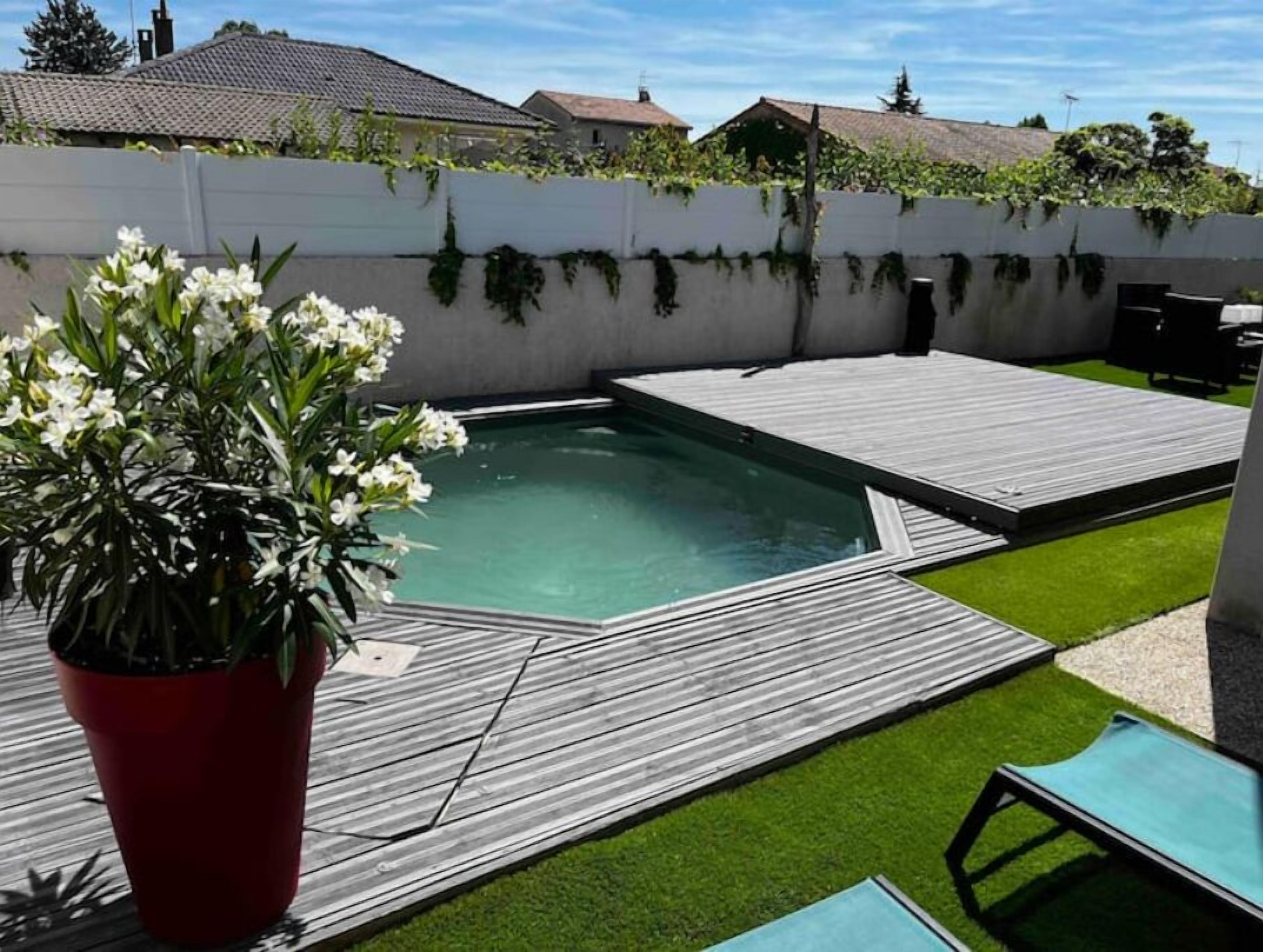 A swimming pool with a wooden deck, surrounded by artificial grass and a white wall.
