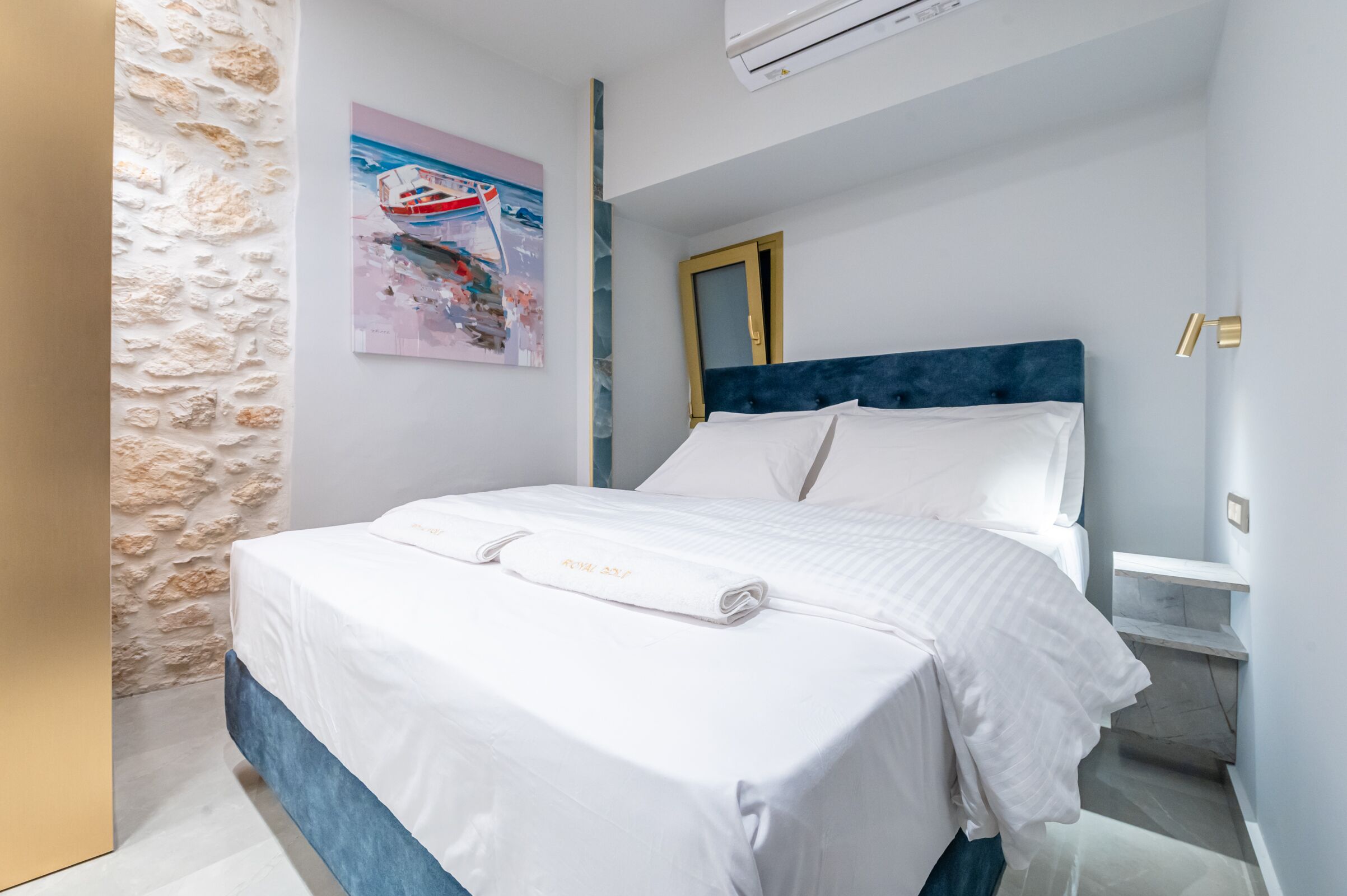 Una cama bien hecha con sábanas y toalla blancos, una pared de piedra, un cuadro de un barco y una lámpara moderna.