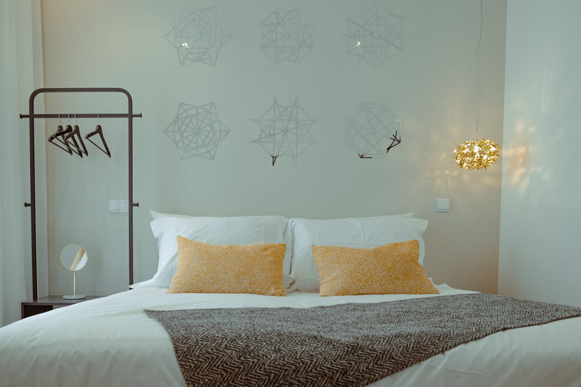 A bedroom with a bed, two pillows, a bedside table, a wall-mounted light fixture, and geometric wall art.