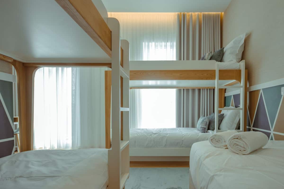 Ein Schlafraum mit Etagenbett, zwei Betten, einem Fenster mit Vorhängen und einem hölzernen Kopfteil.