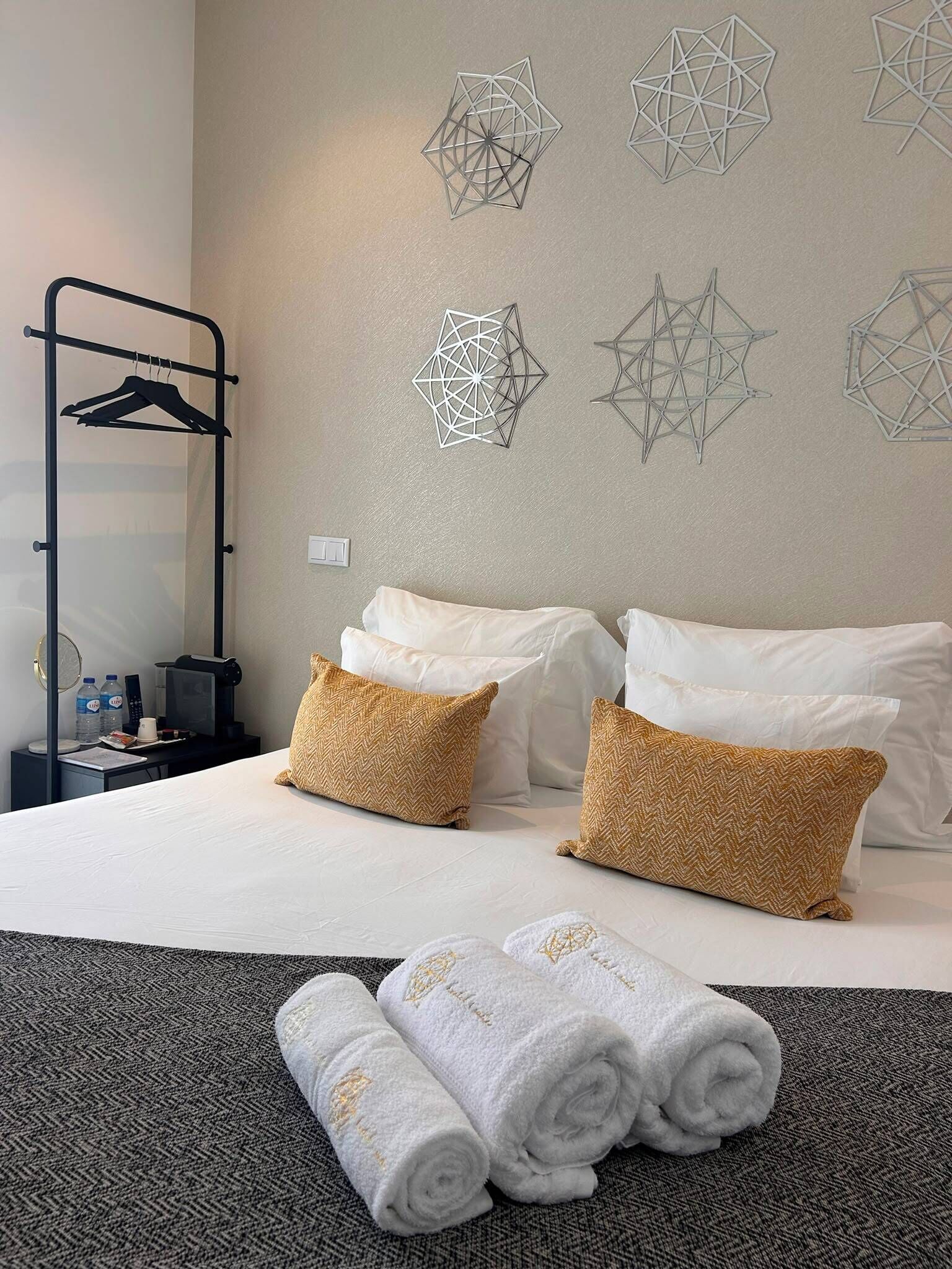 A hotel room with a bed, two pillows, a towel, a nightstand, a water bottle, a remote, and a wall art piece.