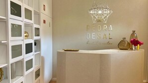 A reception desk with a geometric logo, a white shelving unit with glass doors, and a wooden floor. - Pedra de Sal Hostel & Suites (Rio Maior)