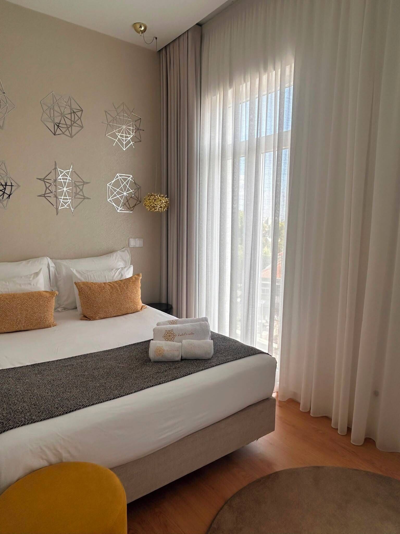 A modern hotel room with a large bed, a small round table, and a view of the city through sheer curtains.