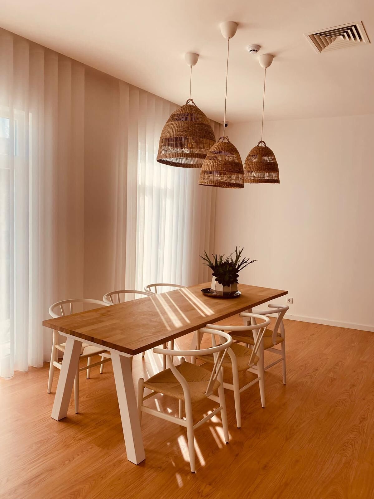 Een eethoek met een houten tafel en witte stoelen, drie rieten hanglampen en een potplant op de tafel.