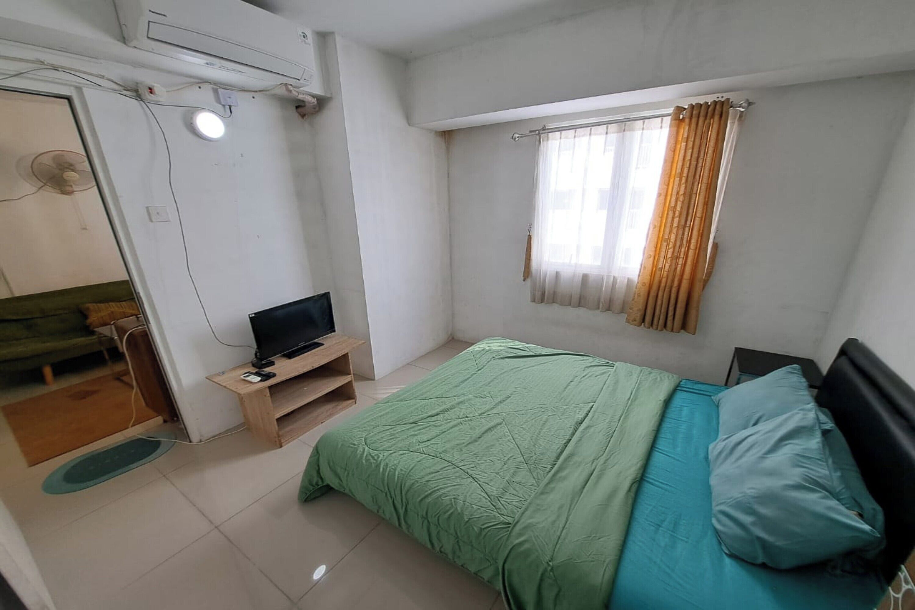 A bedroom with a bed, a TV on a wooden stand, a window with curtains, and a ceiling fan.