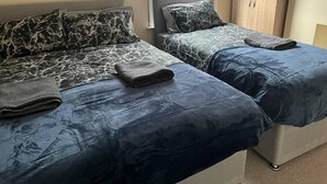 Ein Zimmer mit zwei Betten, jeweils mit dunkelblauer Bettdecke und passenden Kissen. Es gibt ein Fenster mit Jalousien, einen Holzschrank und einen Teppichboden.