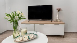 A modern living room with a flat-screen TV on a wooden console, a vase with white flowers, a candle, and a plate with cookies.