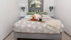 A neatly made bed with a floral bedspread, a bedside table with a lamp, and a window with a view of buildings.