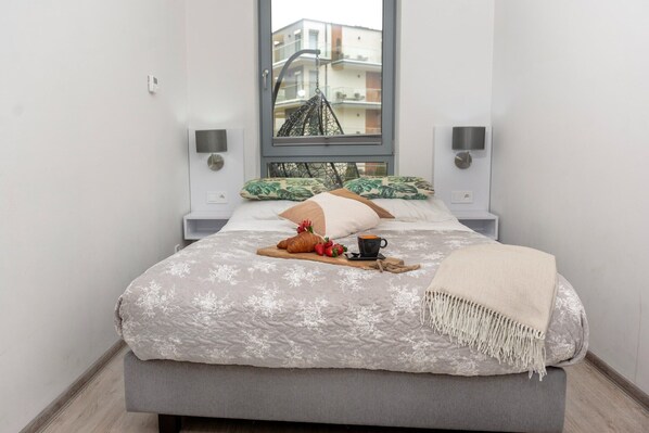 A neatly made bed with a floral bedspread, a bedside table with a lamp, and a window with a view of buildings.