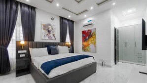 A modern bedroom with a large bed, dark curtains, a flat-screen TV, and a wardrobe. - Diamond Apt - Simply awesome (Lekki)