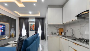 A modern kitchen with white cabinets, a gas stove, and a built-in oven. There is a blue sofa and a small round table with a remote control. - Diamond Apt - Simply awesome (Lekki)