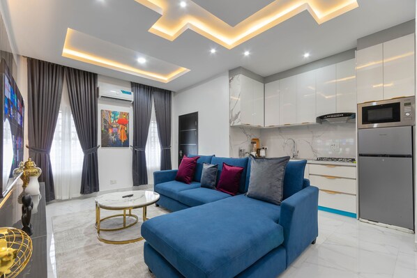 A modern living room with a blue sectional sofa, a white marble coffee table, and a kitchen area with white cabinets and stainless steel appliances. - Diamond Apt - Simply awesome (Lekki)