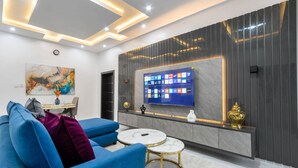 A modern living room with a blue sofa, two marble coffee tables, a large flat-screen TV mounted on a dark wall, and a decorative wall panel. - Diamond Apt - Simply awesome (Lekki)