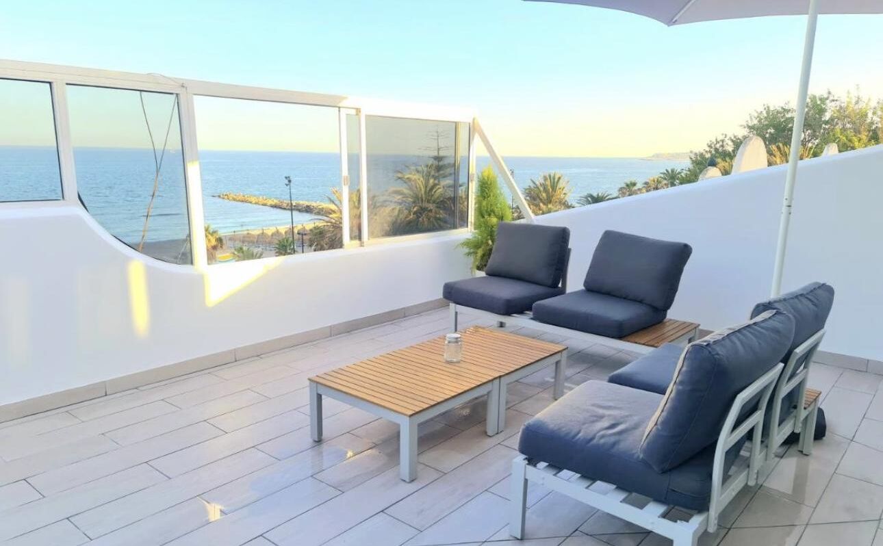 Een balkon met uitzicht op zee, twee blauw gewatteerde stoelen, een houten tafel en een witte balustrade.