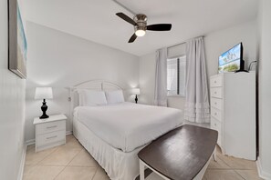 Een slaapkamer met een bed, nachtkastjes, een plafondventilator, een televisie en een raam met gordijnen.